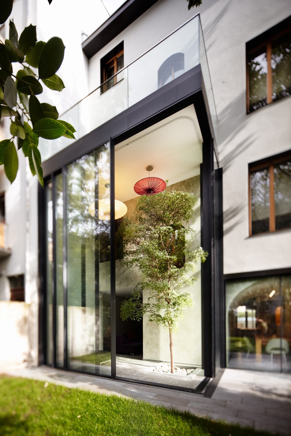 Nowoczesne, przeszklone drzwi przesuwne z czarną ramą, za którymi widać drzewko w minimalistycznym wnętrzu, oświetlone czerwoną lampą.