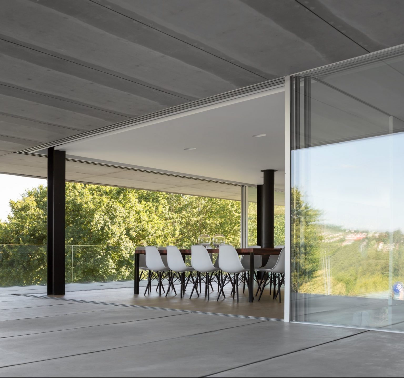 Nowoczesne, przeszklone patio z przesuwnymi drzwiami, betonowa podłoga i sufit, widok na zieleń za oknem, stół z krzesłami w tle.