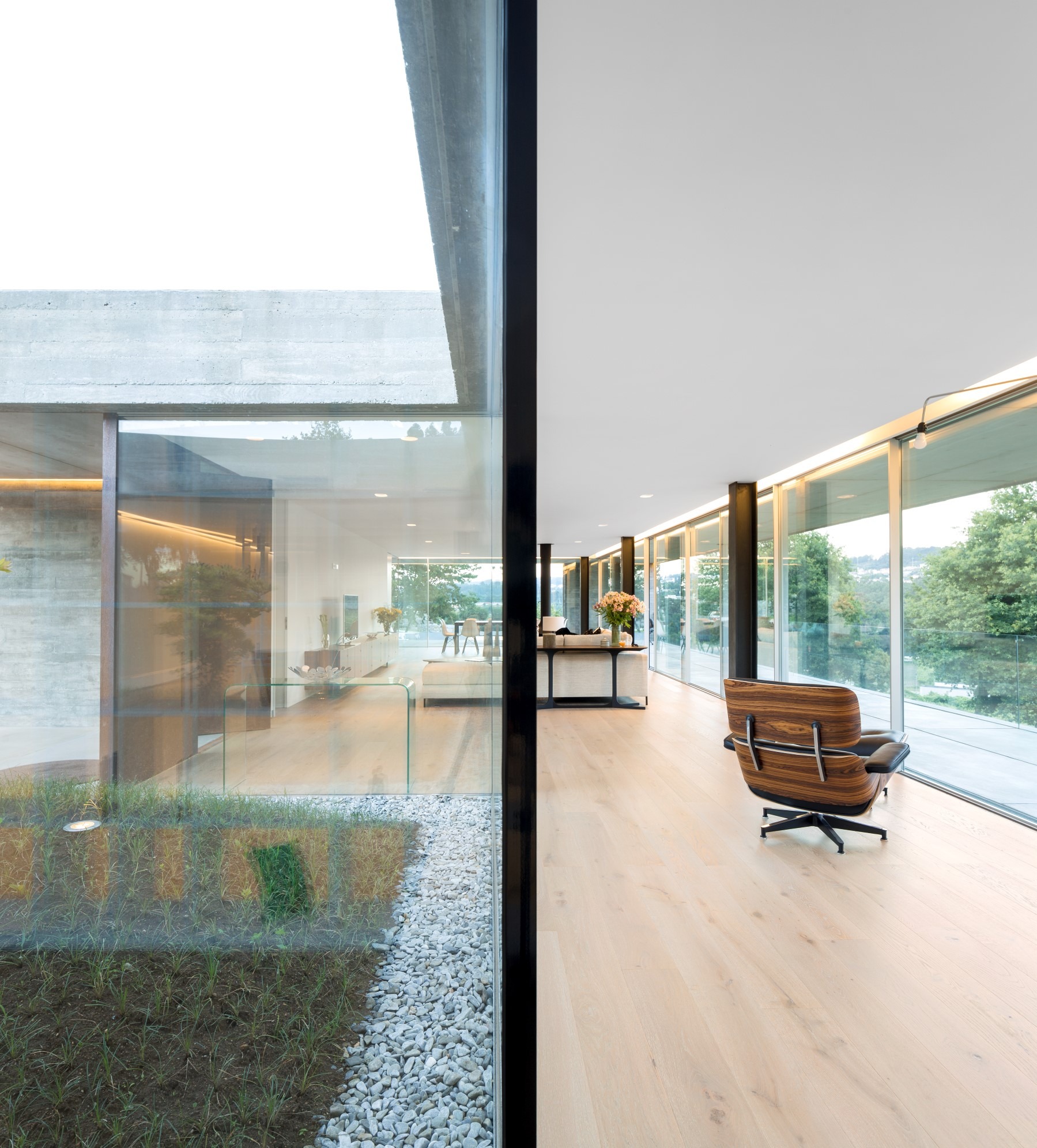 Nowoczesny salon z panoramicznymi oknami przesuwnymi, widok na ogród z kamienną rabatą, fotel Eames Lounge Chair na jasnej drewnianej podłodze.