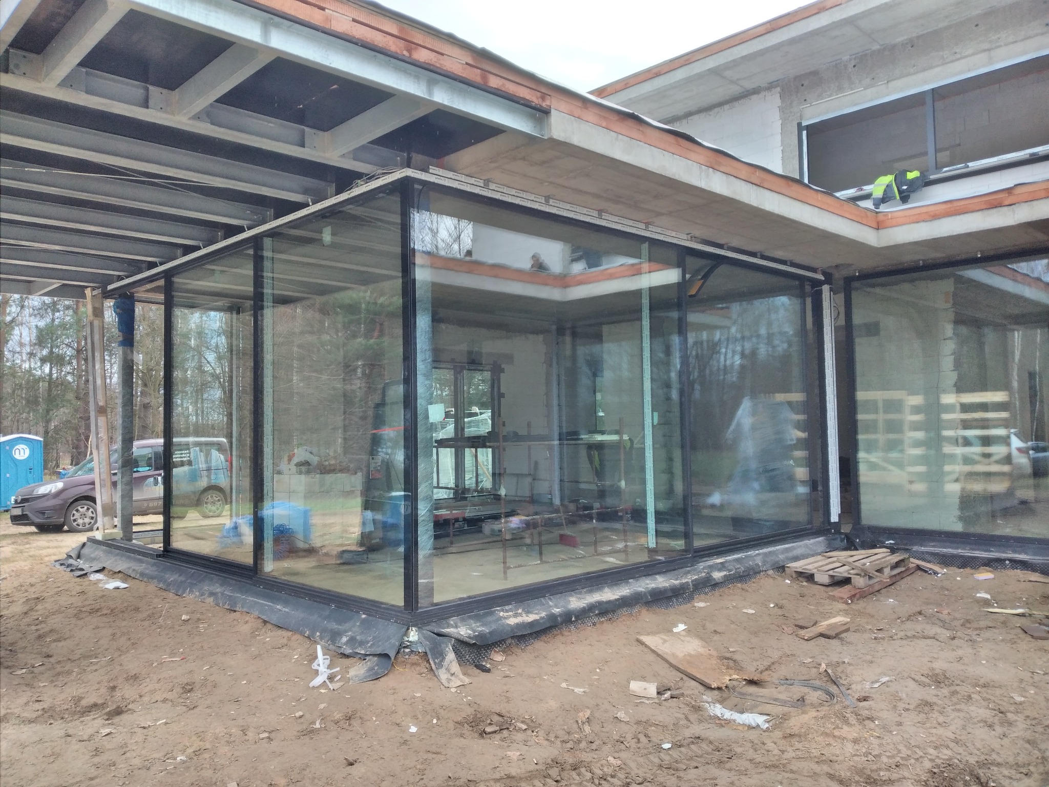Montaż przeszklonej konstrukcji z czarnymi ramami na surowym etapie budowy domu. Widoczne rusztowania wewnątrz i pracownik na dachu. Na zewnątrz zaparkowany samochód i toaleta przenośna.