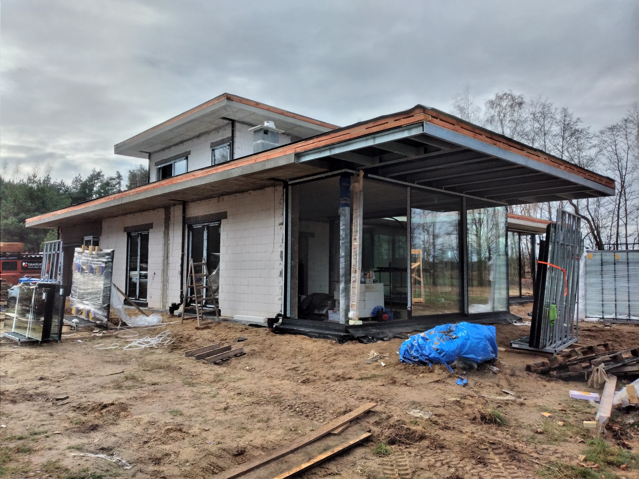 Surowy stan budowy domu jednorodzinnego z dużymi przeszkleniami, widoczne elementy konstrukcyjne dachu i rusztowania, na pierwszym planie teren budowy z piaskiem i materiałami budowlanymi.