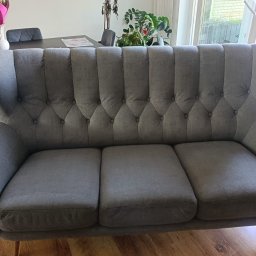 Clean Protection Katarzyna Hejno - Szara sofa trzyosobowa z guzikami na oparciu, na drewnianej podłodze, w tle stół z krzesłami i bukiet kwiatów.