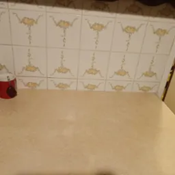 Beżowy blat kuchenny z widocznymi płytkami ceramicznymi w kwiatowe wzory, czerwony kubek z zawieszką stoi na blacie przy krawędzi kuchenki