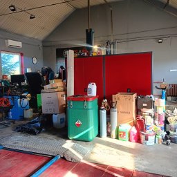 Clean Protection Katarzyna Hejno - Magazyn z różnorodnymi przedmiotami w Lublinie: puszki z farbami, pojemniki na chemikalia, butle gazowe, kartony i dywan, wszystko zgromadzone w pomieszczeniu z czerwonymi szafkami i oknem w tle.