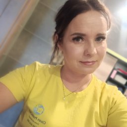 Clean Protection Katarzyna Hejno - Portret kobiety w żółtej koszulce z logo firmy sprzątającej, uśmiechnięta, z delikatnym makijażem, w tle fragment wnętrza z półkami.