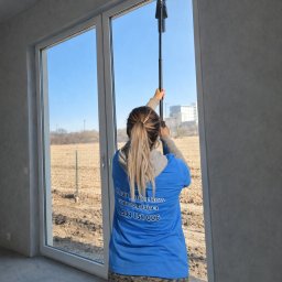 Clean Protection Katarzyna Hejno - Kobieta w niebieskiej koszulce z logo firmy sprzątającej myje okno w nowym budynku za pomocą profesjonalnej myjki teleskopowej. Widok zza pleców, jasne wnętrze.