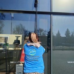 Clean Protection Katarzyna Hejno - Pracownica w Wólce Abramowickiej myje okna na zewnątrz budynku, używając specjalistycznego sprzętu teleskopowego. Widoczny budynek i otoczenie.