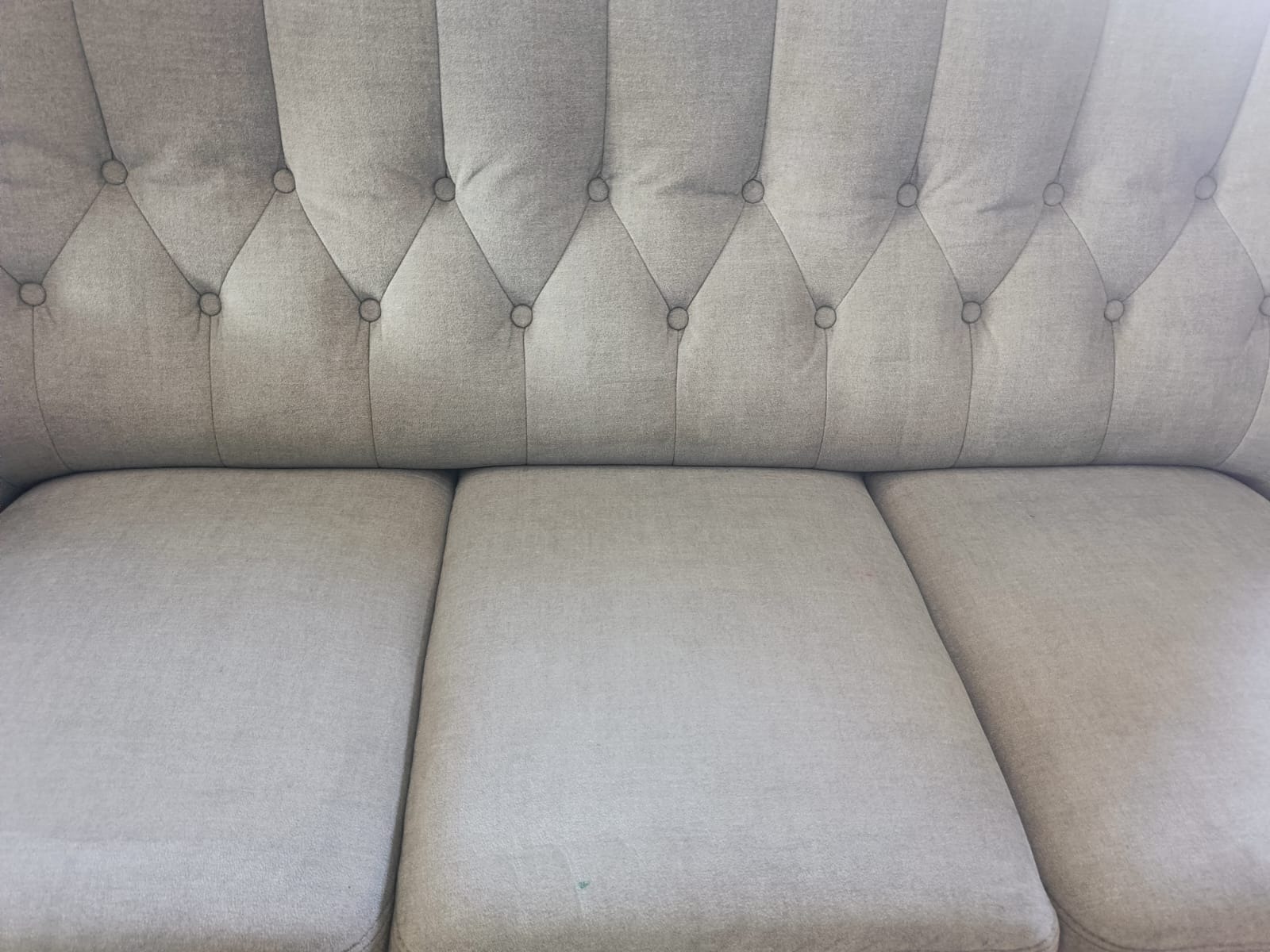 Fragment sofy z jasną tapicerką, widoczne pikowania z guzikami na oparciu i trzy siedziska, delikatne plamy na środkowym siedzisku.