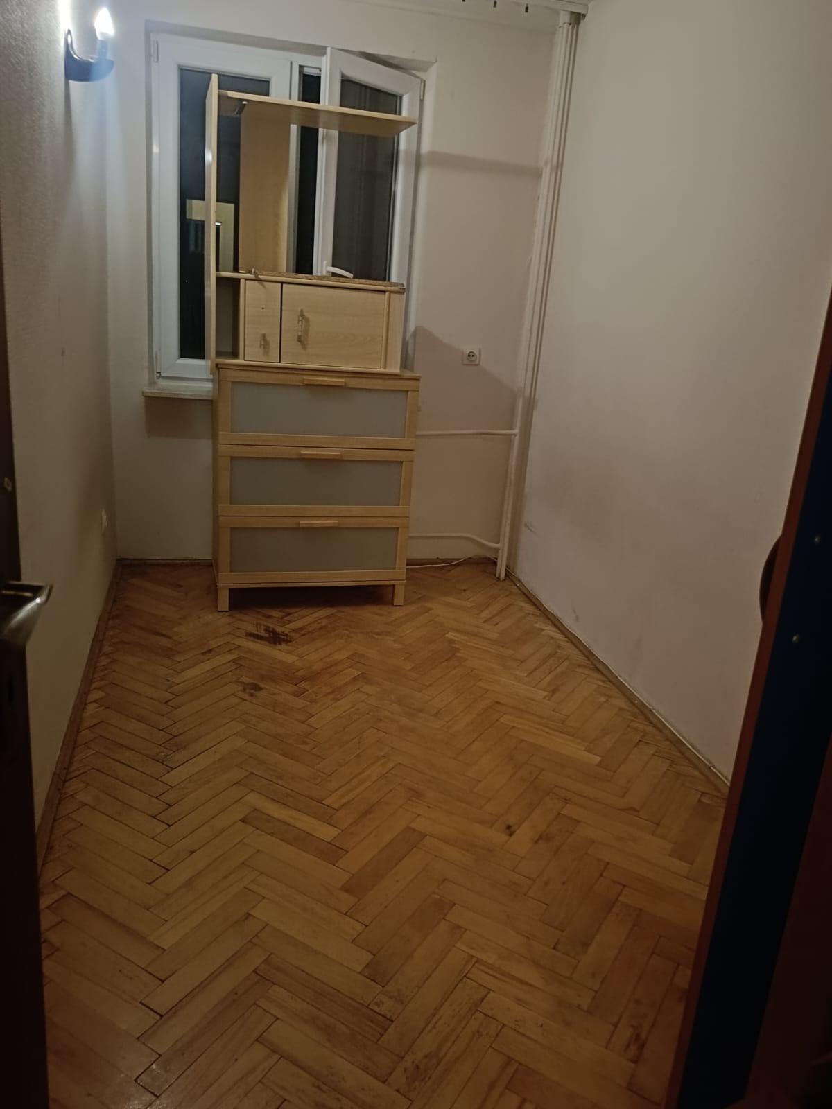 Pusty pokój z parkietem w jodełkę i drewnianą komodą z nadstawką przy oknie.