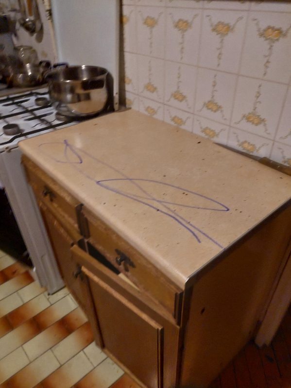 Blat kuchenny z brązowego lastryko, zabrudzony niebieskim flamastrem, widoczne otwarte drzwiczki szafki i fragment kuchenki gazowej z garnkami w tle.