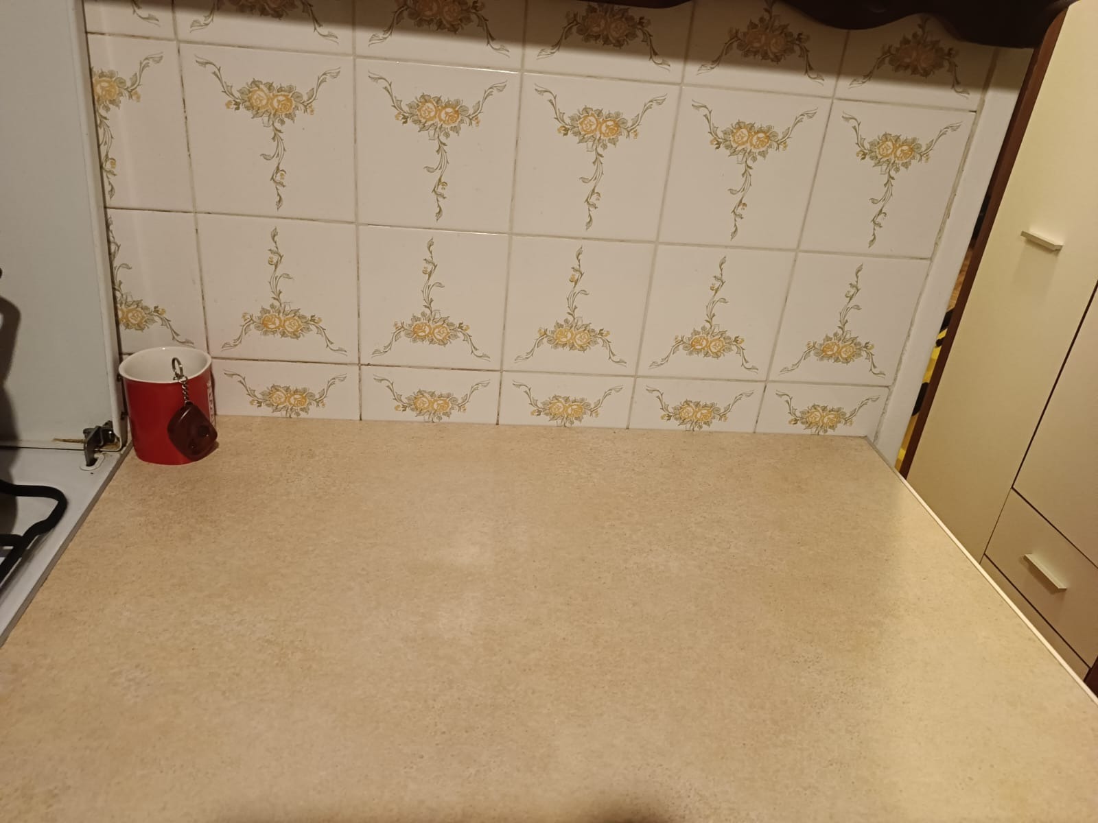 Beżowy blat kuchenny z widocznymi płytkami ceramicznymi w kwiatowe wzory, czerwony kubek z zawieszką stoi na blacie przy krawędzi kuchenki