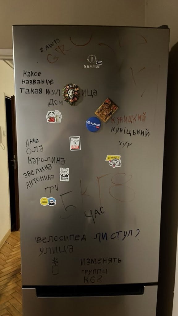 Frontalny widok na brudną, srebrną lodówkę z naklejkami i napisami wykonanymi markerem, stojącą w mieszkaniu.