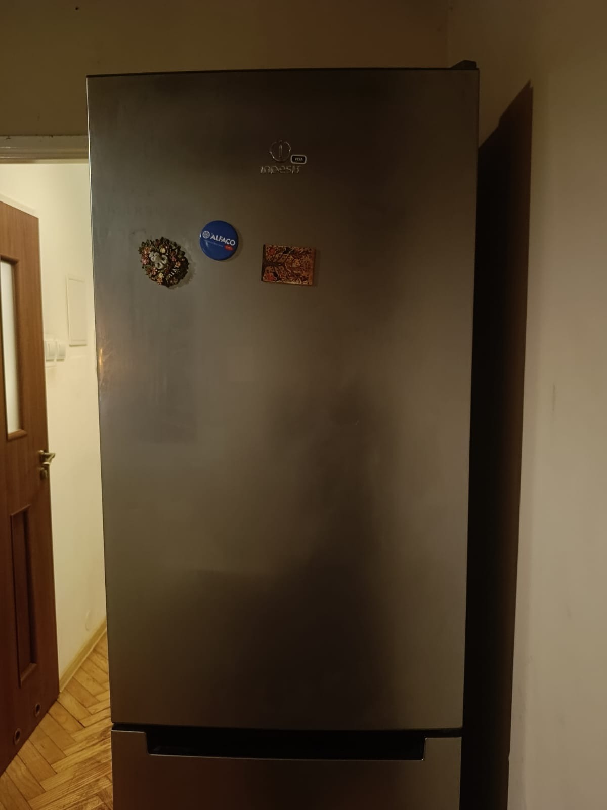 Stalowa lodówka z magnesami na drzwiach, w tle widoczny fragment drzwi i ściana.