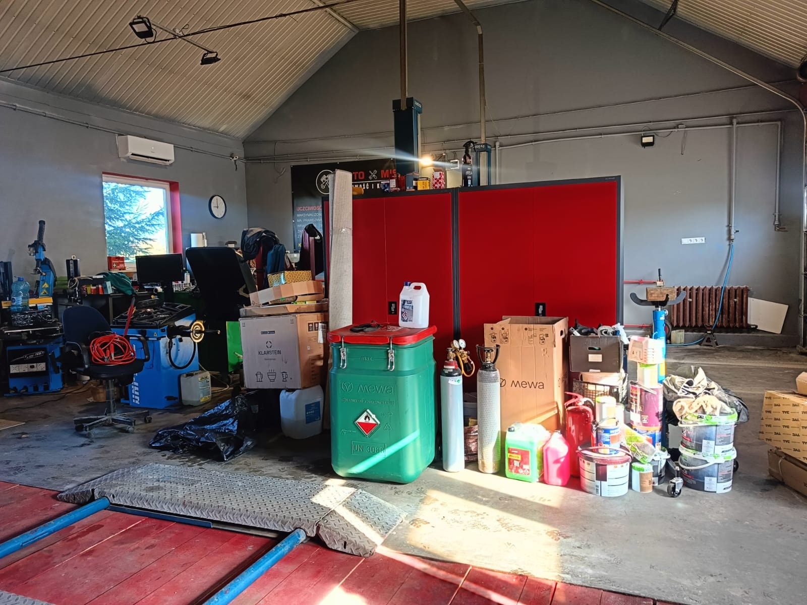 Magazyn z różnorodnymi przedmiotami w Lublinie: puszki z farbami, pojemniki na chemikalia, butle gazowe, kartony i dywan, wszystko zgromadzone w pomieszczeniu z czerwonymi szafkami i oknem w tle.