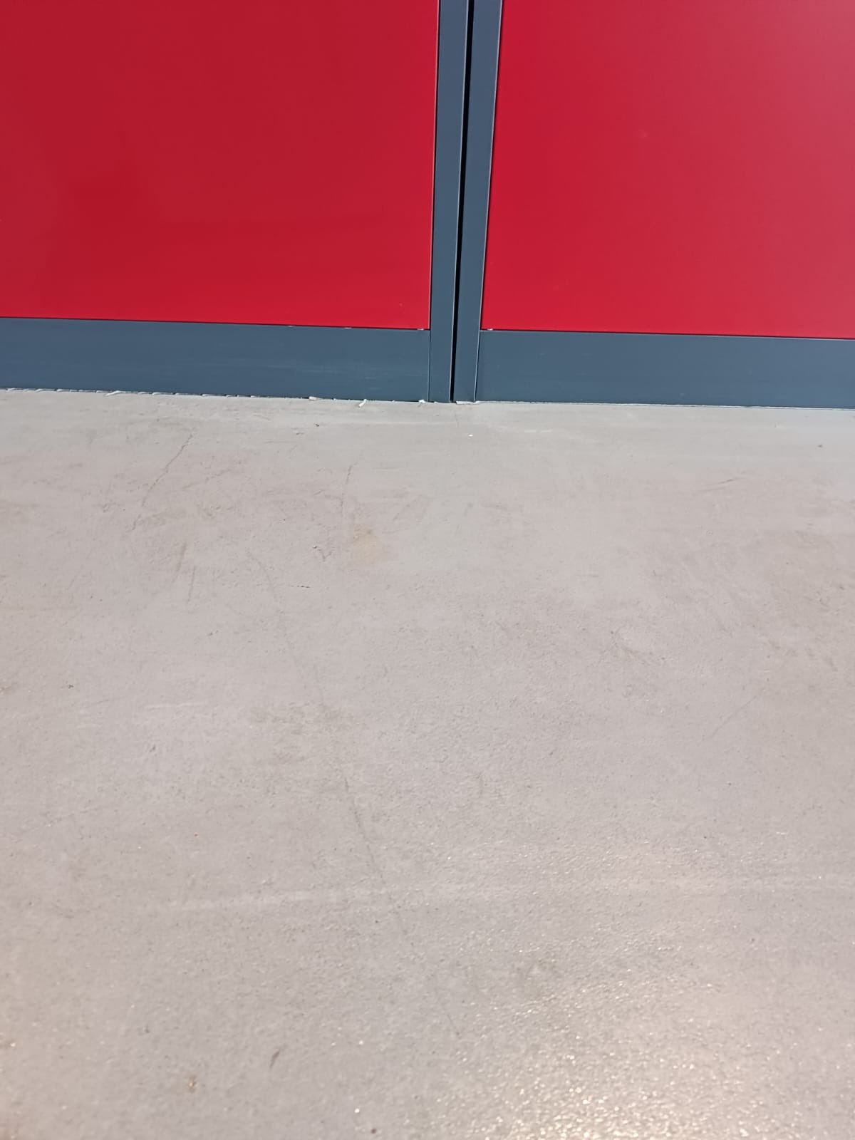 Szara betonowa posadzka z widocznymi rysami i śladami, dolna część czerwonej ściany z szarym cokołem.