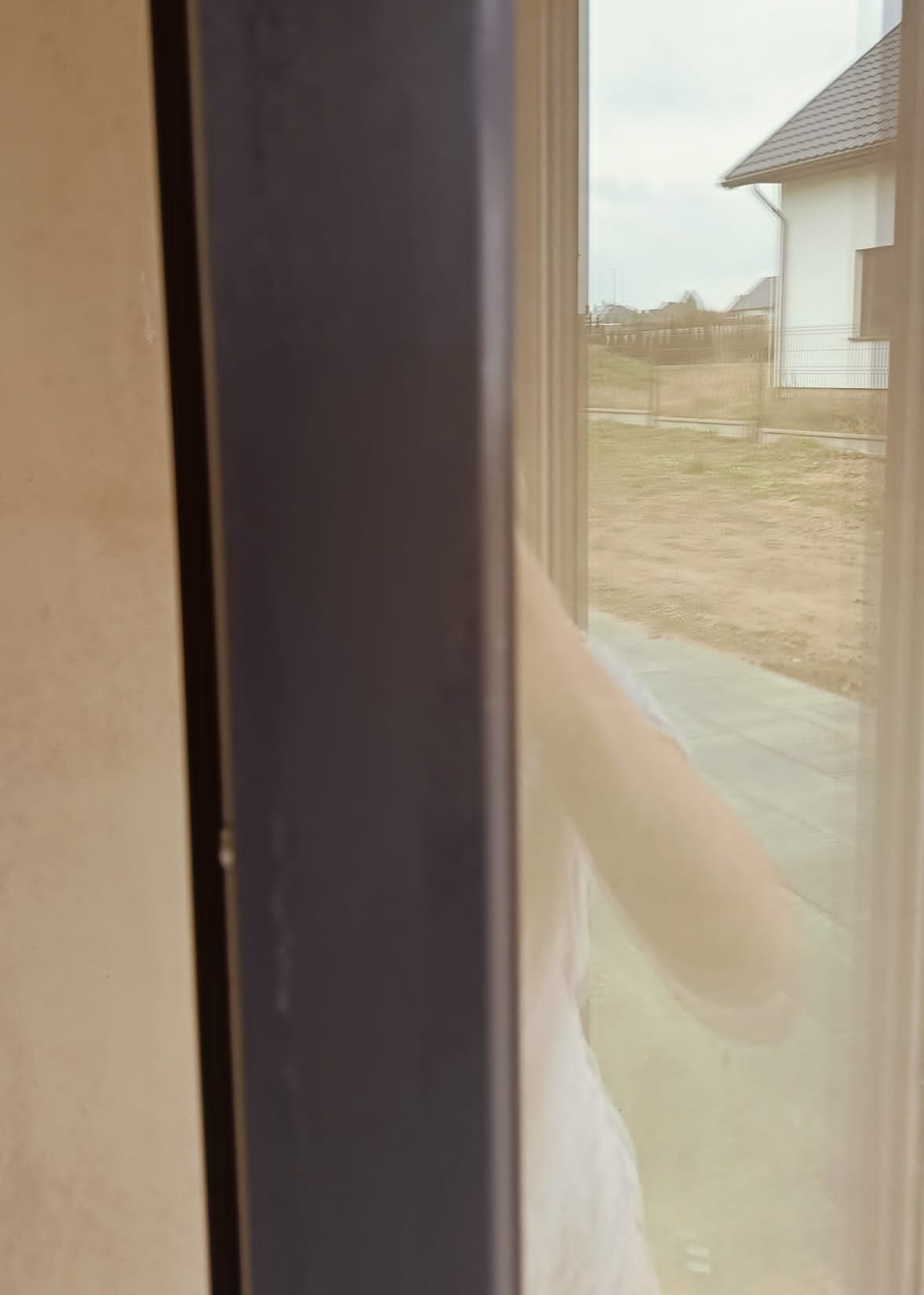 Osoba w białej koszulce myje okno, widok z wnętrza domu na ogród i dom sąsiada przez częściowo umyte szkło.