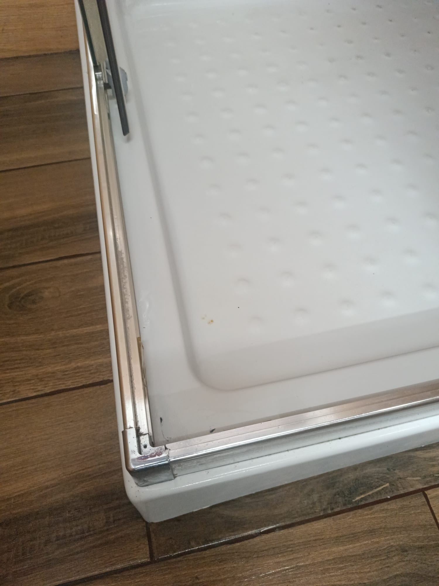 Biały brodzik kabiny prysznicowej z aluminiowymi profilami, widoczne ślady użytkowania i osady na ramie, ujęcie z bliska na tle drewnopodobnych płytek podłogowych.