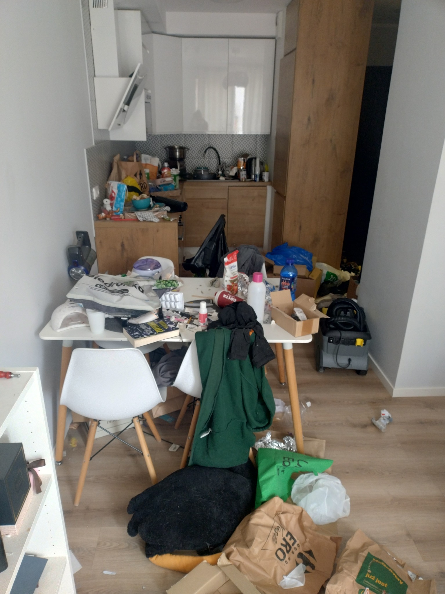 Zabałaganiona kuchnia z widocznym stołem zawalonym przedmiotami, torbami i sprzętem AGD. Na podłodze leżą worki i śmieci. Widok na wnętrze przed sprzątaniem.