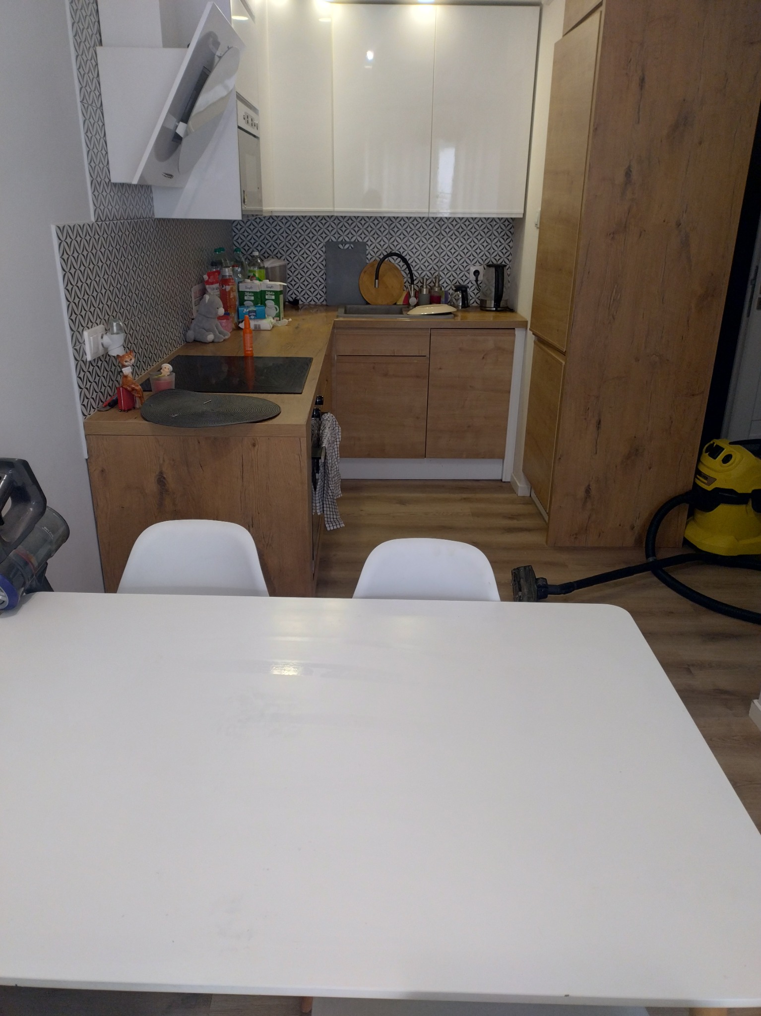 Ujęcie na kuchnię z białymi szafkami, drewnianymi blatami i białym stołem. W tle odkurzacz. Widoczny fragment odkurzonej podłogi.