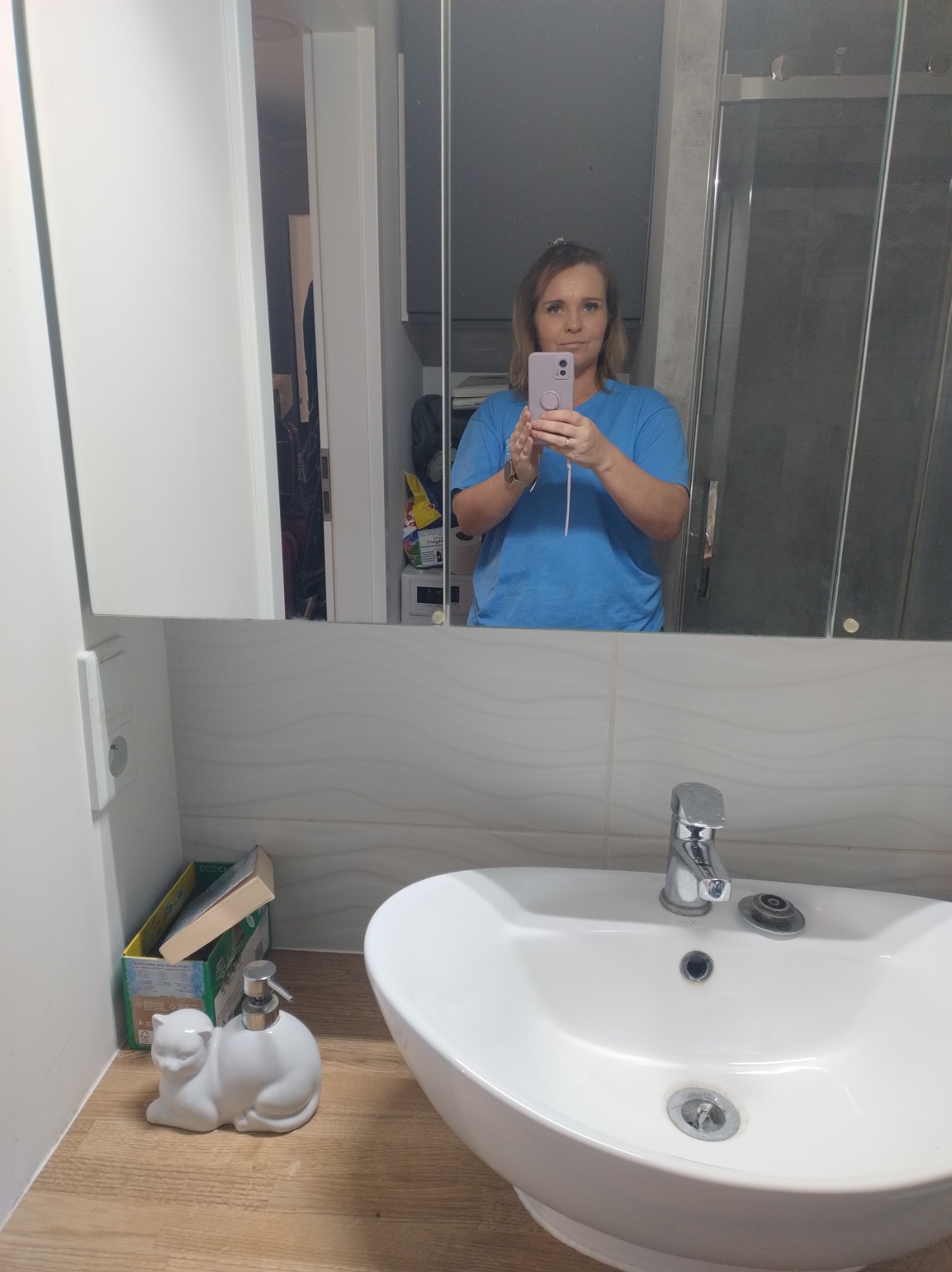 Osoba w niebieskiej koszulce robi selfie w lustrze łazienki. Widoczna umywalka, dozownik mydła w kształcie kota i szafka z akcesoriami w tle.