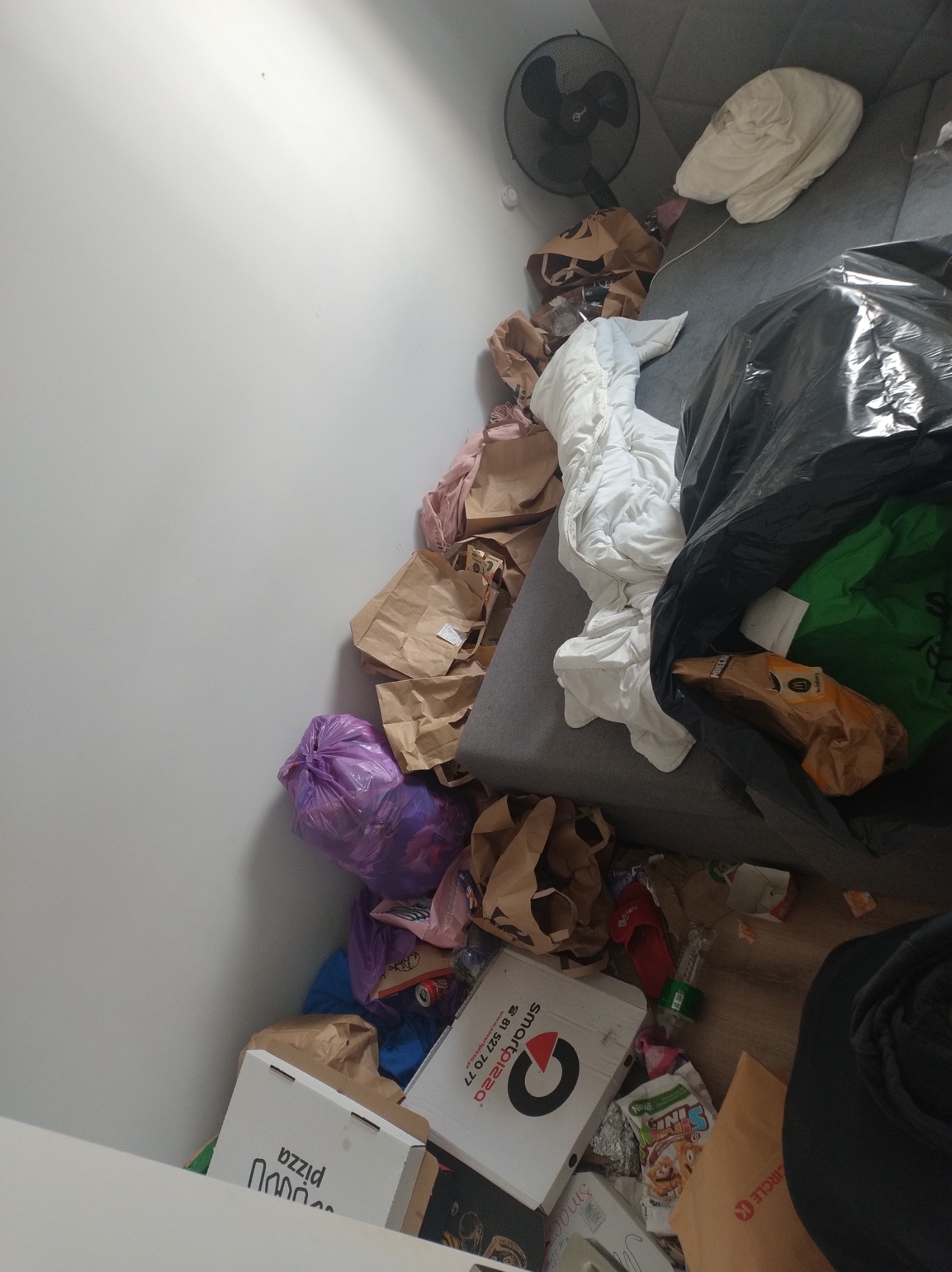 Zabrudzone mieszkanie z workami śmieci, kartonami po pizzy, torbami i innymi odpadami na podłodze i kanapie. Widok z góry na bałagan.