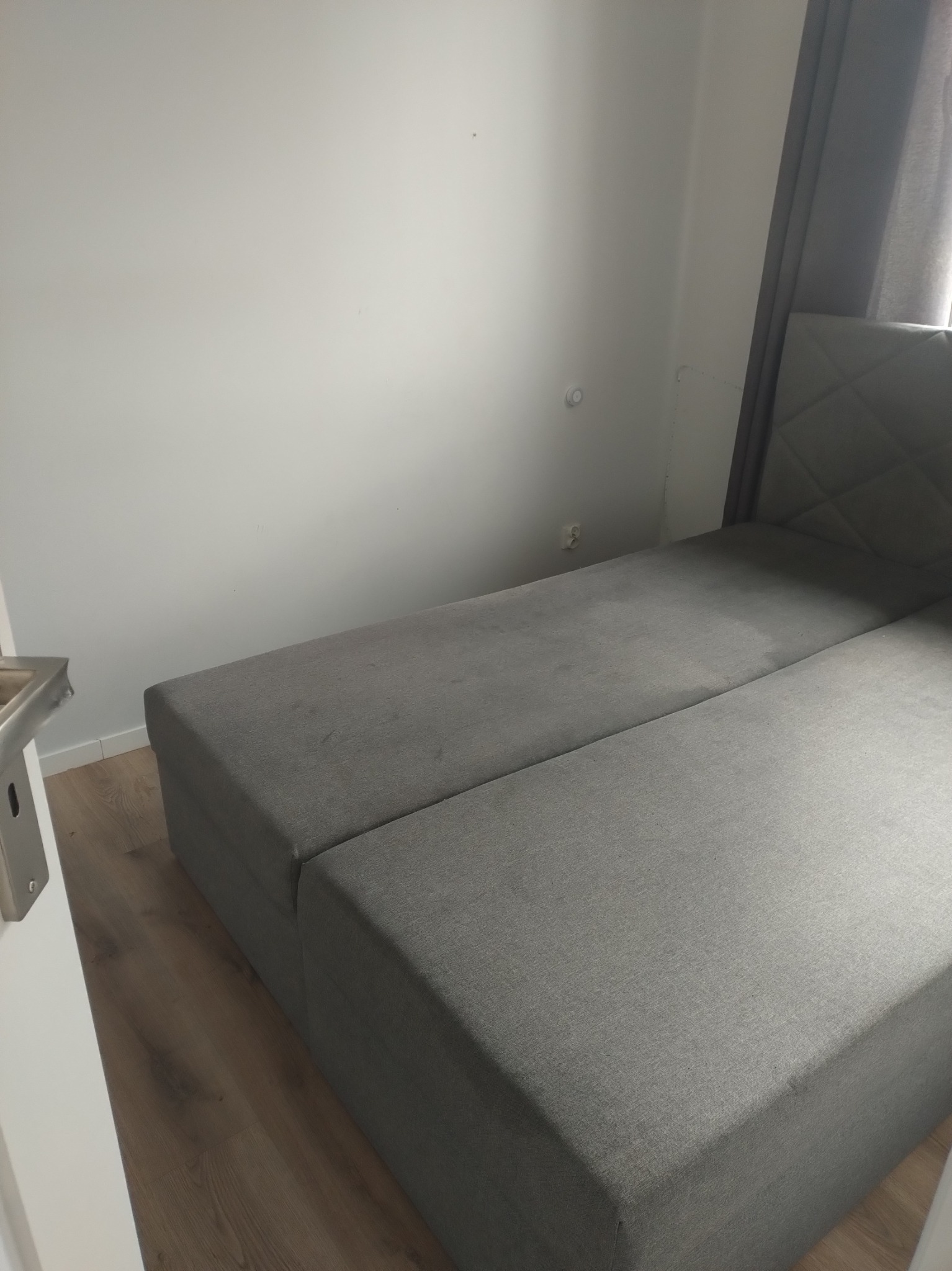 Szary narożnik z zagłówkiem w pokoju po sprzątaniu, widoczne gniazdko elektryczne i fragment zasłony. Czyste ściany i podłoga.