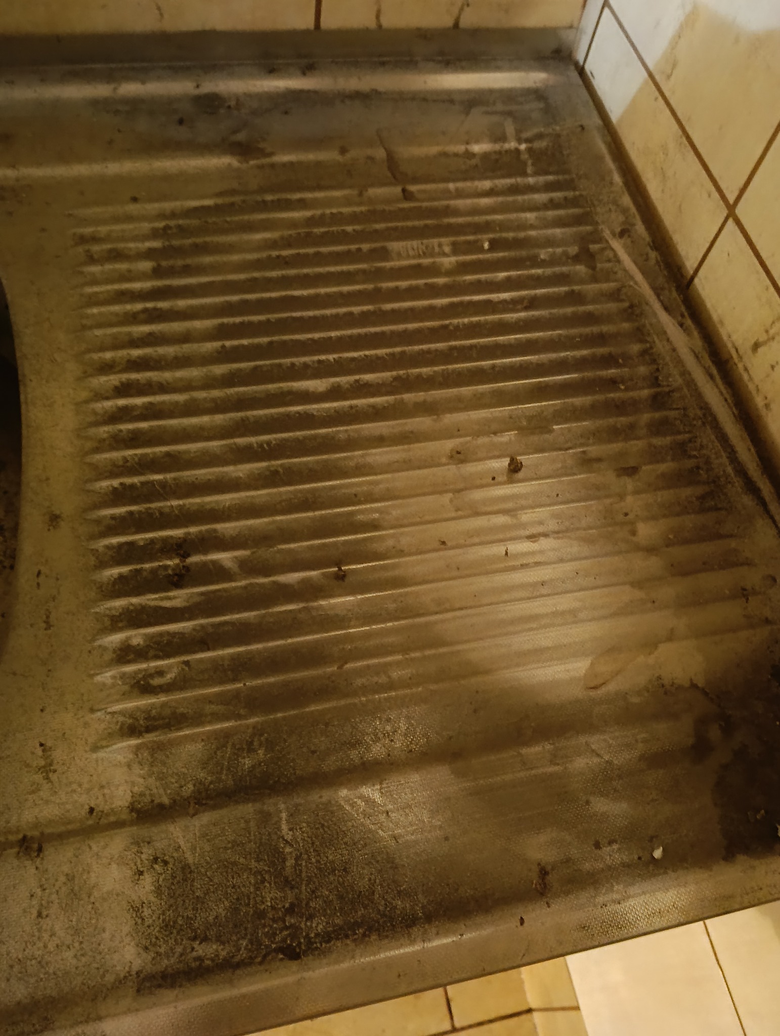 Zabrudzona, metalowa taca o żłobkowanej powierzchni, przylegająca do ściany z płytek. Widoczne plamy i osady, sugerujące potrzebę gruntownego czyszczenia.