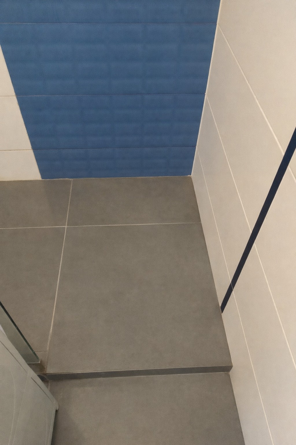 Wykończona łazienka z szarymi płytkami podłogowymi i biało-niebieskimi płytkami ściennymi. Widoczny fragment kabiny prysznicowej. Minimalistyczny design.