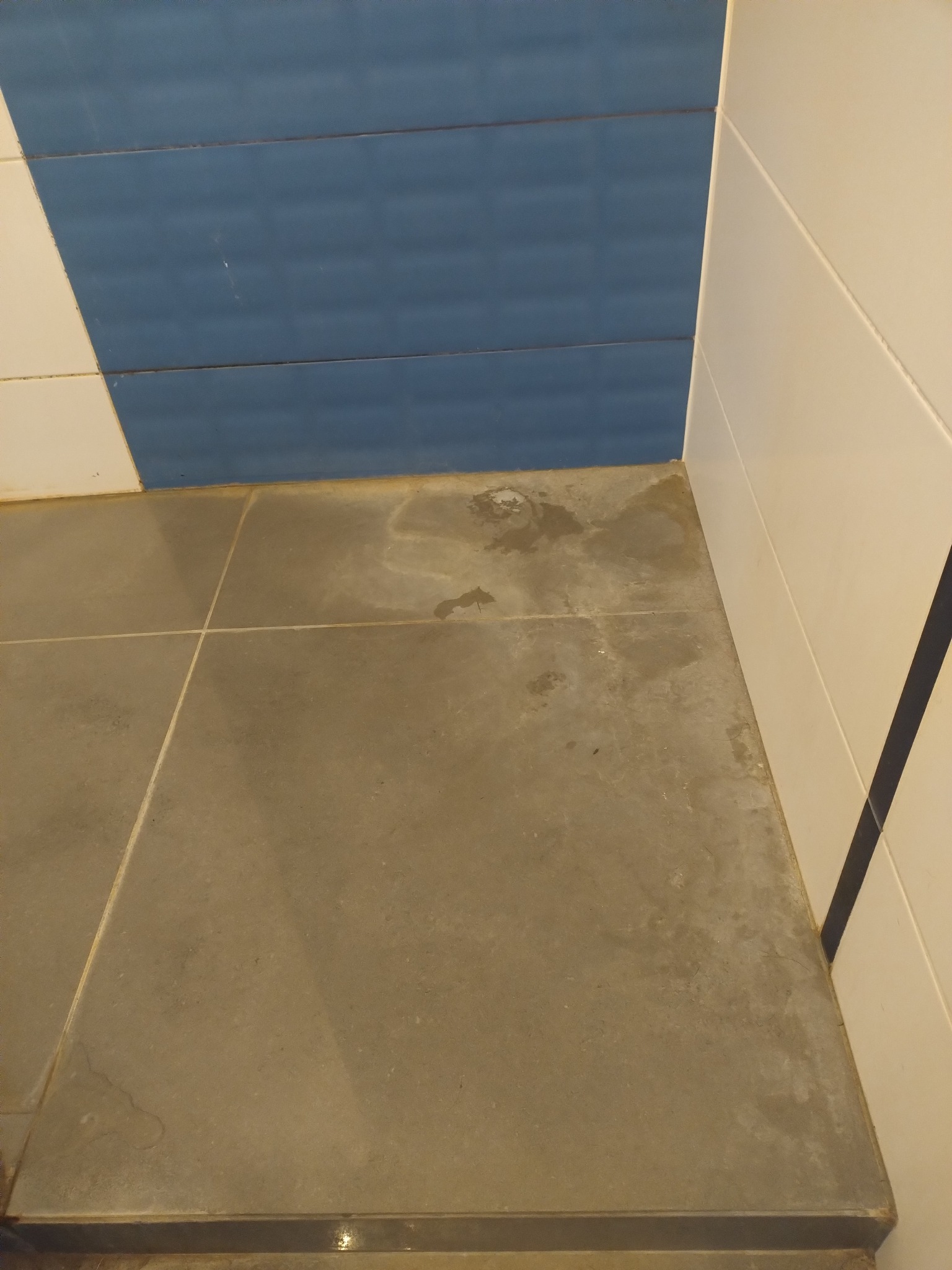 Brudna, szara podłoga z płytek w łazience, widoczne zacieki i plamy, biało-niebieskie płytki na ścianie w tle. Perspektywa z góry, ukazująca potrzebę gruntownego sprzątania.