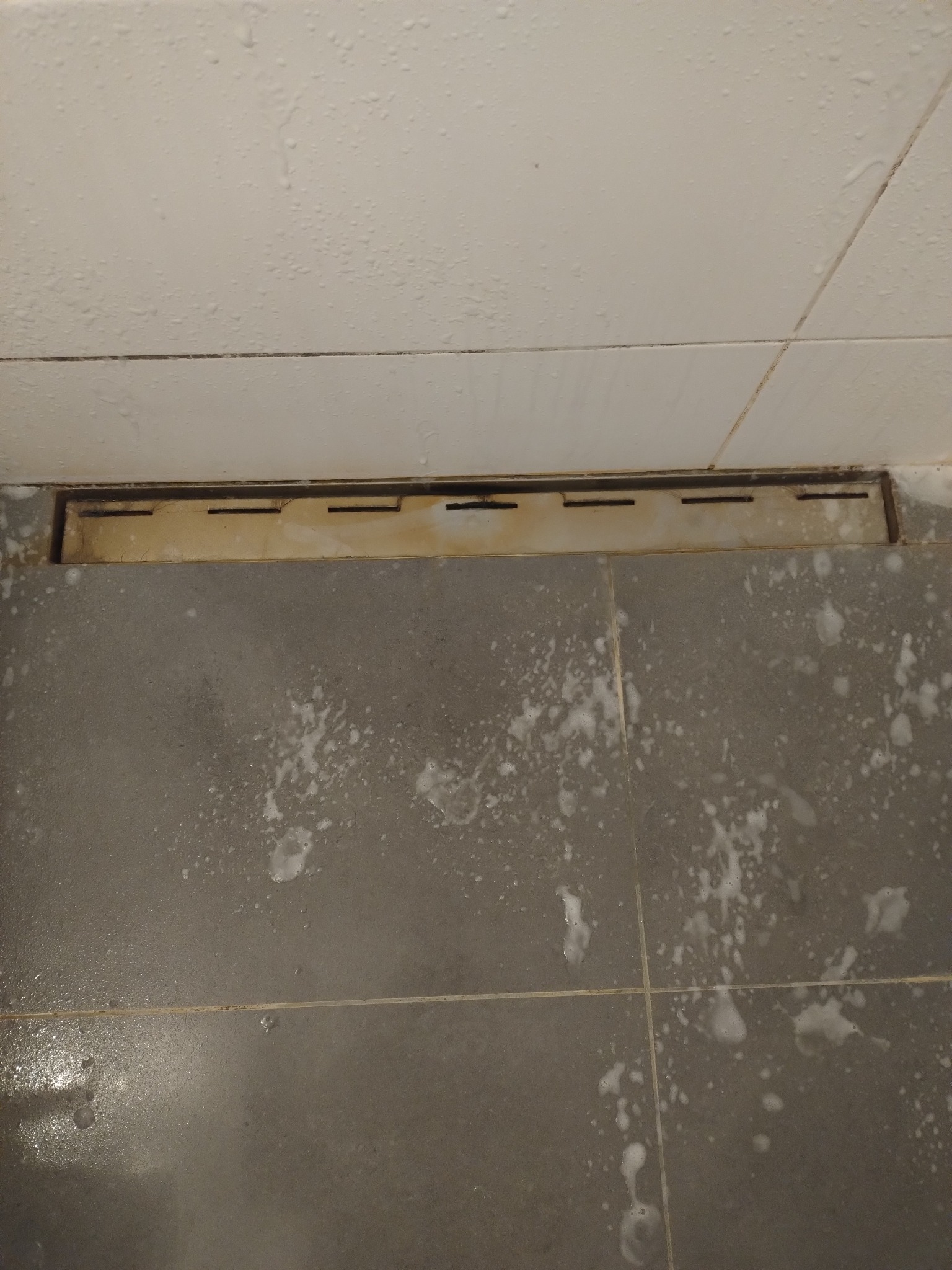 Szare płytki podłogowe w łazience, pokryte pianą czyszczącą, z widocznym odpływem liniowym. Świeżo po aplikacji środka czyszczącego, przed spłukaniem.