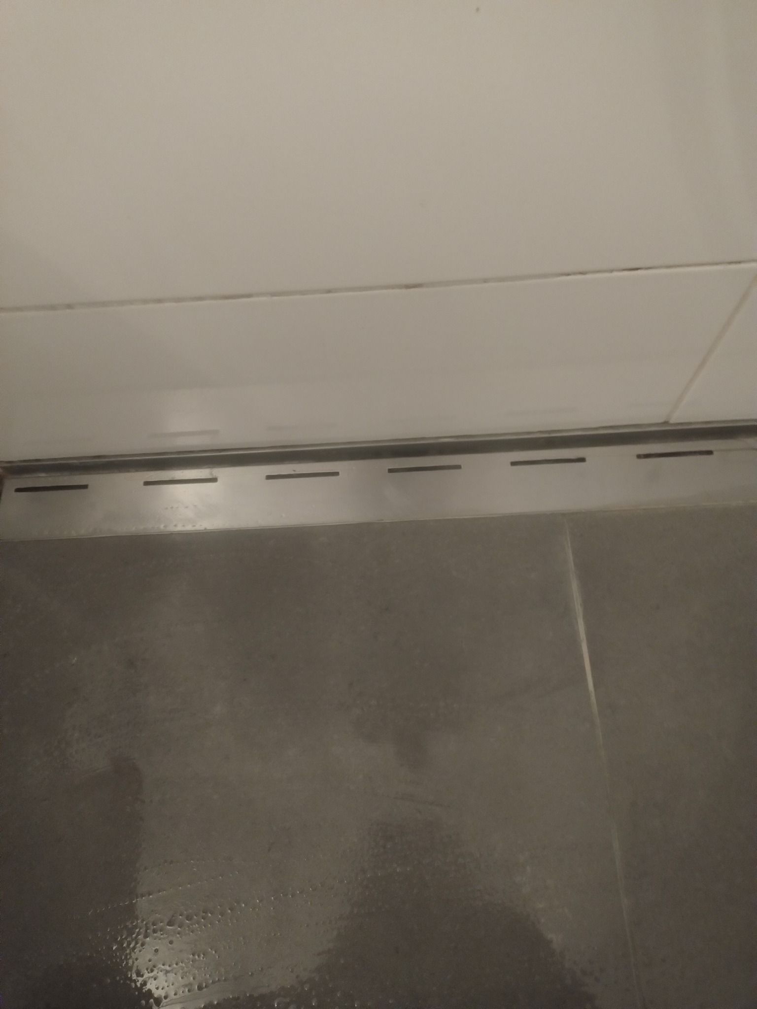 Szczegółowe ujęcie czystej, szarej podłogi z odpływem liniowym w łazience, po sprzątaniu. Widoczne płytki i fugi bez zabrudzeń, z delikatnymi śladami wilgoci.