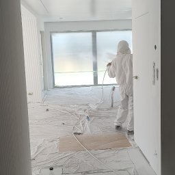 USŁUGI REMONTOWO-BUDOWLANE IDEAL HAUS MARIUSZ WITEK - Malowanie natryskowe ścian w Lublinie: Pracownik w kombinezonie ochronnym maluje na biało ściany w pomieszczeniu, zabezpieczonym folią i kartonem, widoczny agregat malarski i wiadro z farbą Flugger.