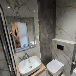 USŁUGI REMONTOWO-BUDOWLANE IDEAL HAUS MARIUSZ WITEK - Nowoczesna łazienka z marmurowymi płytkami, wiszącą toaletą i umywalką nablatową. W lustrze odbija się fragment remontowanego pomieszczenia.