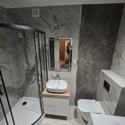 USŁUGI REMONTOWO-BUDOWLANE IDEAL HAUS MARIUSZ WITEK - Nowoczesna łazienka z kabiną prysznicową typu walk-in, umywalką na szafce z drewnianym blatem i toaletą podwieszaną. Marmurowe płytki na ścianach i podłodze.