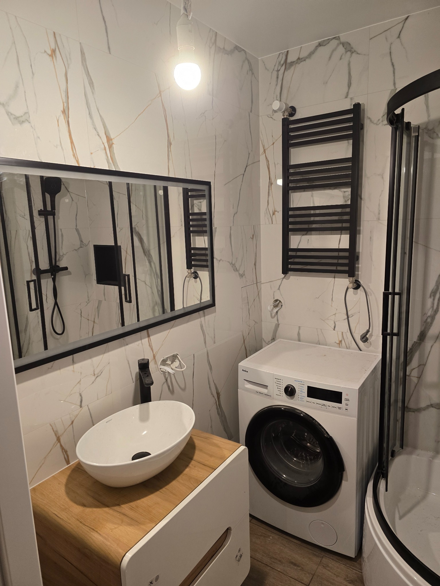 Nowoczesna łazienka z marmurowymi płytkami, owalną umywalką na drewnianej szafce, czarną armaturą i grzejnikiem drabinkowym obok pralki.