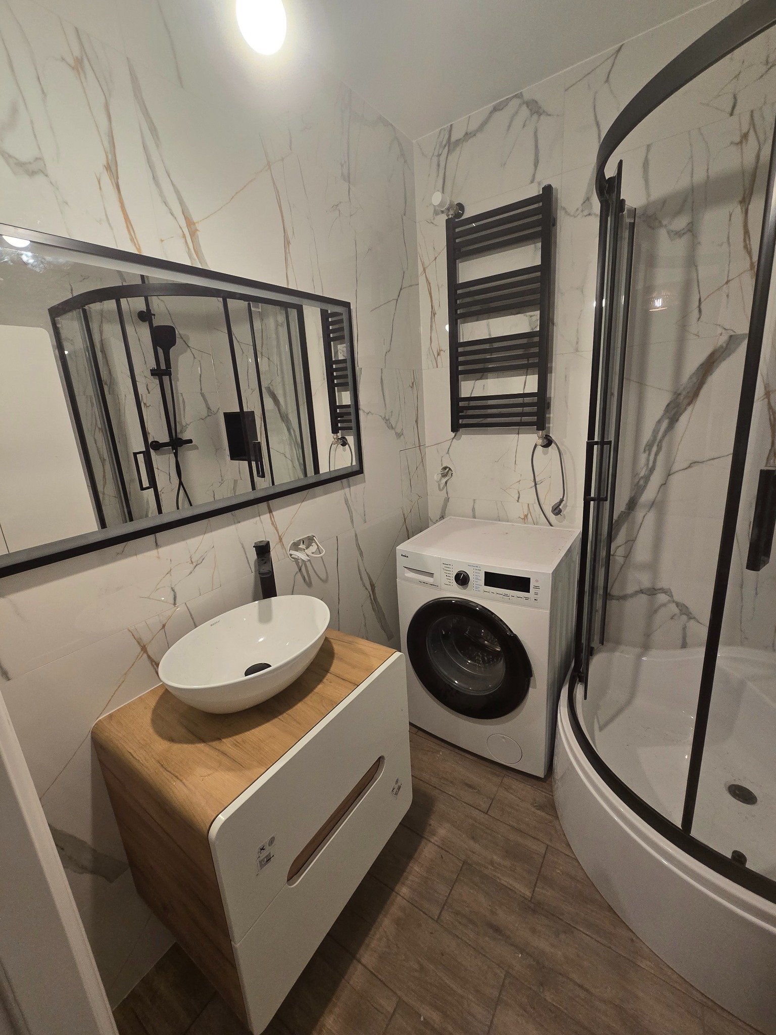 Nowoczesna łazienka po remoncie: białe marmurowe płytki, umywalka nablatowa na drewnianej szafce, czarna rama lustra i kabiny prysznicowej, pralka.