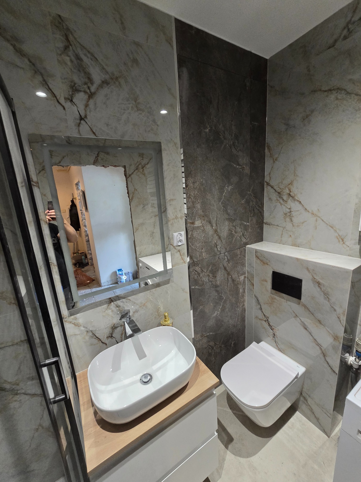 Nowoczesna łazienka z marmurowymi płytkami, wiszącą toaletą i umywalką nablatową. W lustrze odbija się fragment remontowanego pomieszczenia.