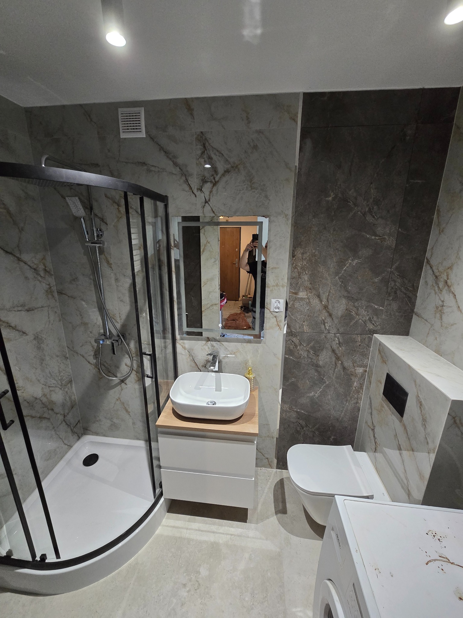 Nowoczesna łazienka z kabiną prysznicową typu walk-in, umywalką na szafce z drewnianym blatem i toaletą podwieszaną. Marmurowe płytki na ścianach i podłodze.