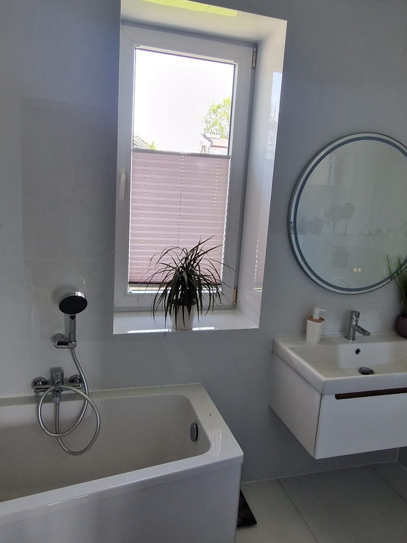 Jasna łazienka z wanną, umywalką i lustrem. Na parapecie okna roślina doniczkowa. Minimalistyczny wystrój, białe płytki i armatura. Nowoczesne rolety w oknie.