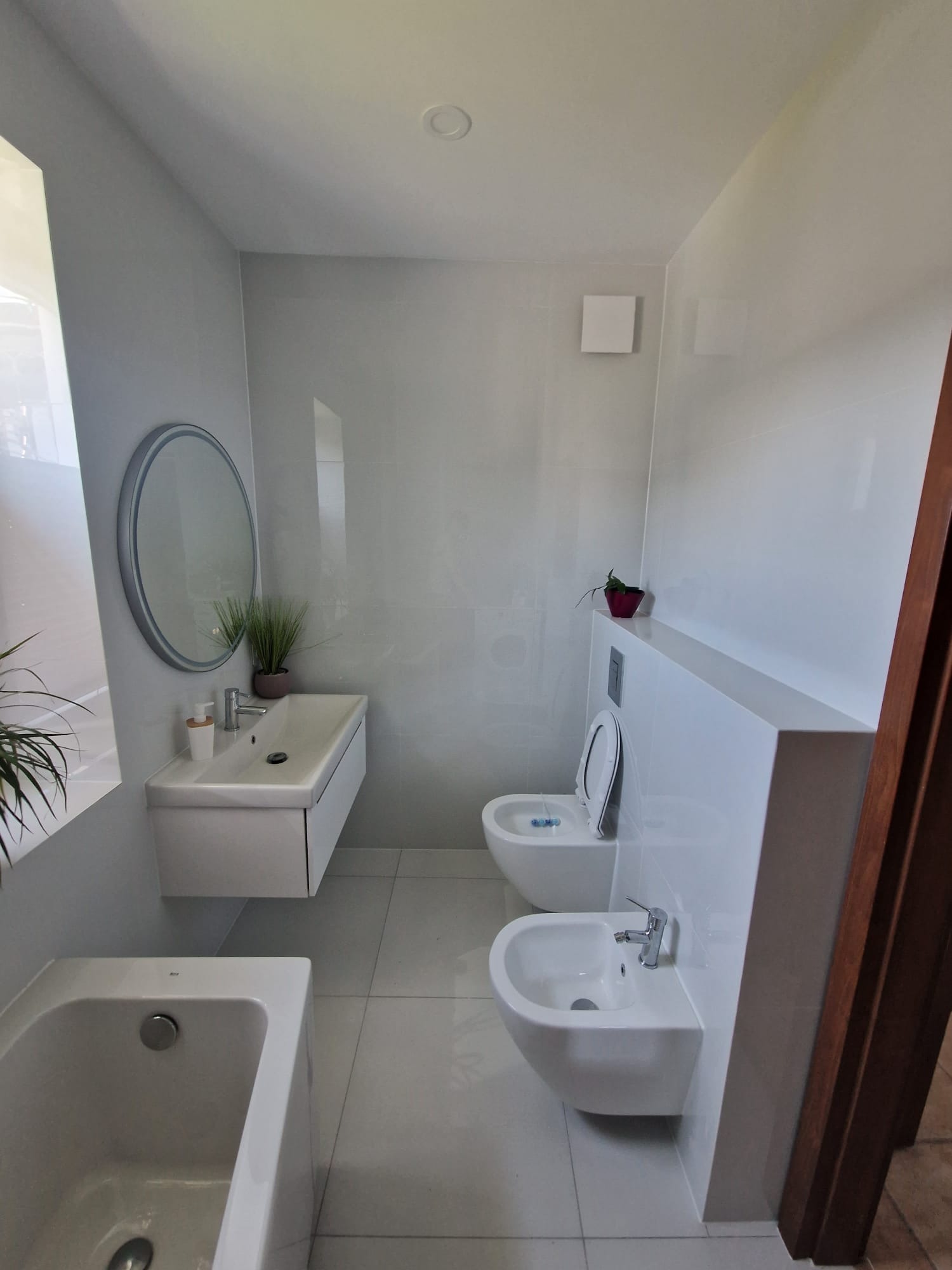 Jasna łazienka z białą armaturą: umywalka z szafką, okrągłe lustro, toaleta podwieszana i bidet. Minimalistyczny design z roślinami jako akcentami dekoracyjnymi.