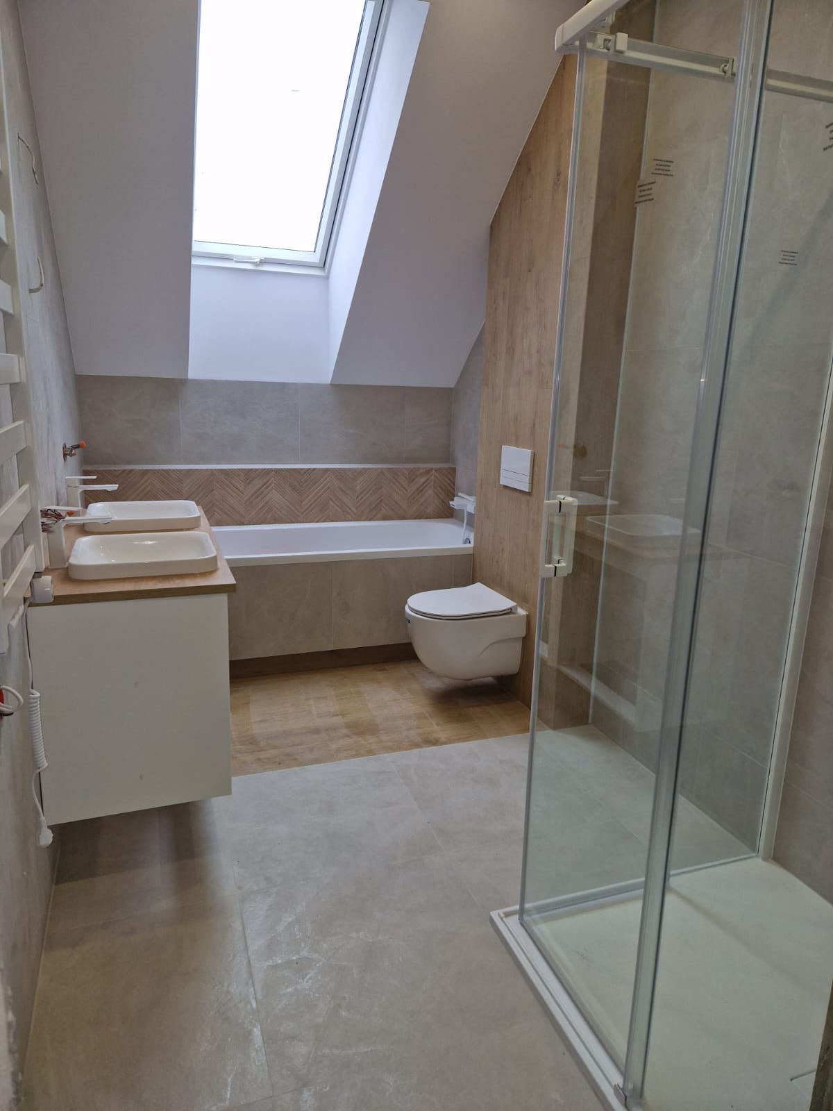 Jasna łazienka na poddaszu z oknem dachowym. Umywalka z szafką, toaleta podwieszana i kabina prysznicowa. Płytki w odcieniach beżu i brązu, wzór jodełki nad wanną.