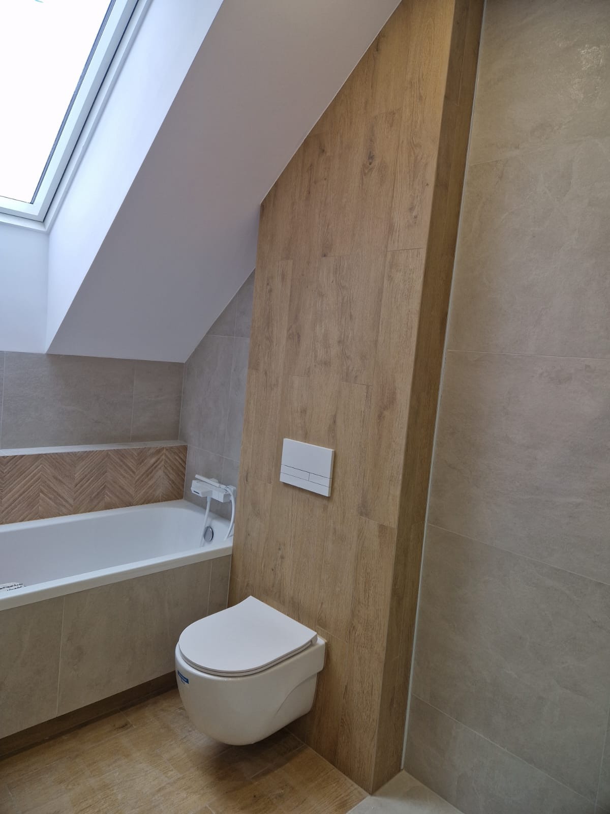 Wykończona łazienka na poddaszu z oknem dachowym. Beżowe płytki, drewnopodobna ściana, podwieszana toaleta i wanna z dekoracyjnym wykończeniem.