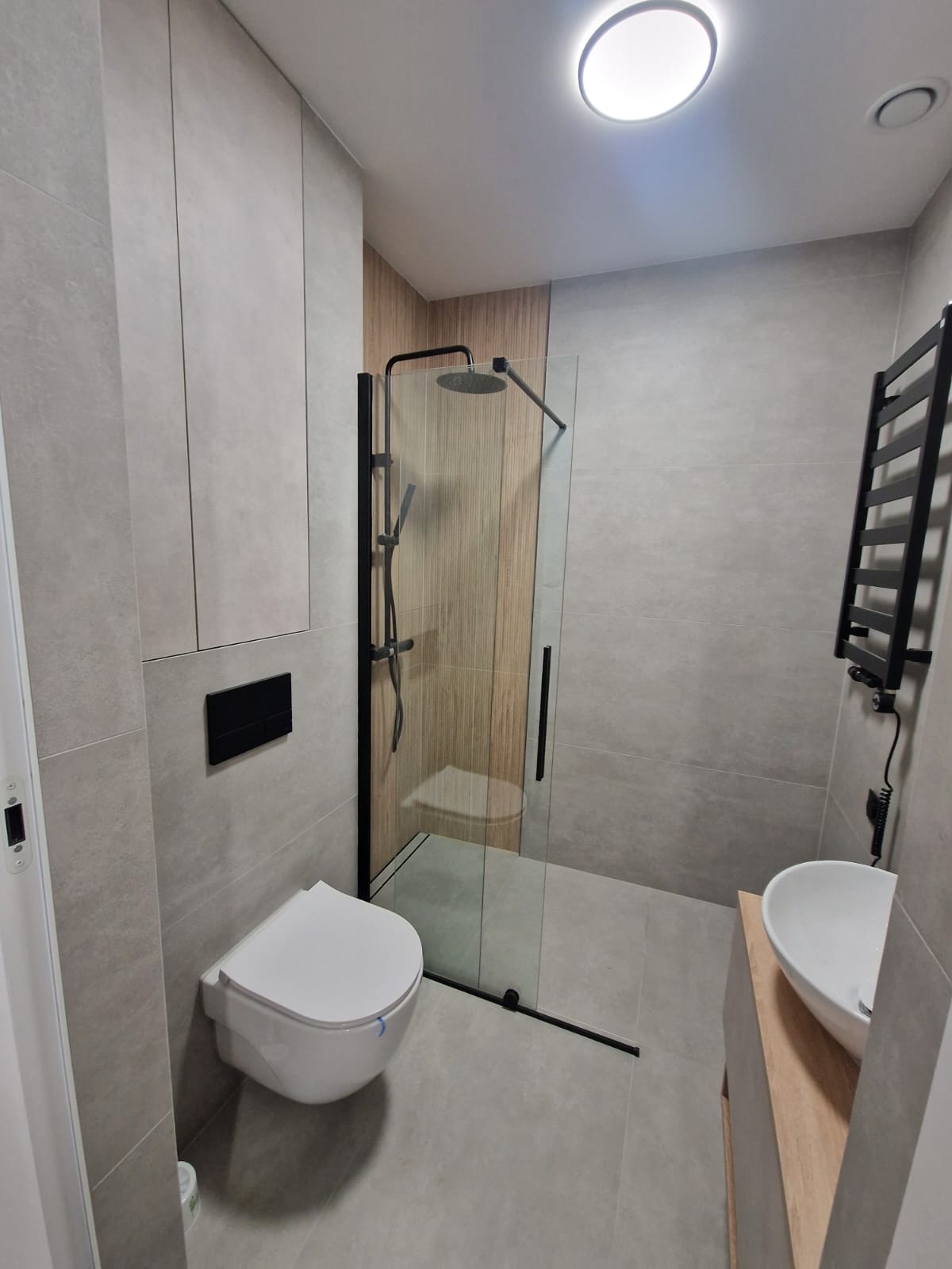 Nowoczesna łazienka w szarościach z kabiną prysznicową typu walk-in z czarnymi profilami, podwieszaną toaletą i grzejnikiem drabinkowym. Minimalistyczny design.