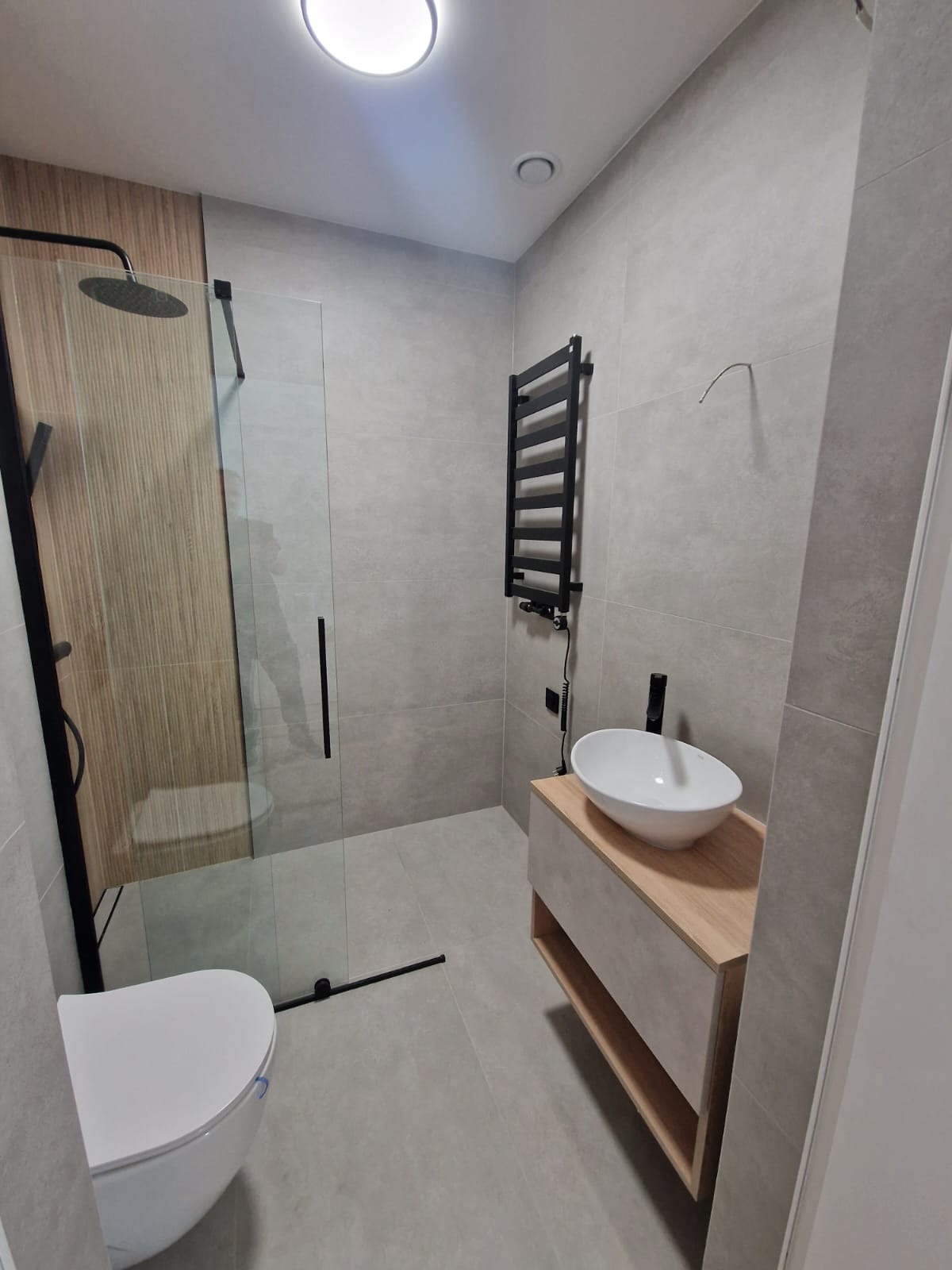 Nowoczesna łazienka z kabiną prysznicową typu walk-in, umywalką nablatową na drewnianej szafce i grzejnikiem drabinkowym w kolorze czarnym na szarych płytkach.