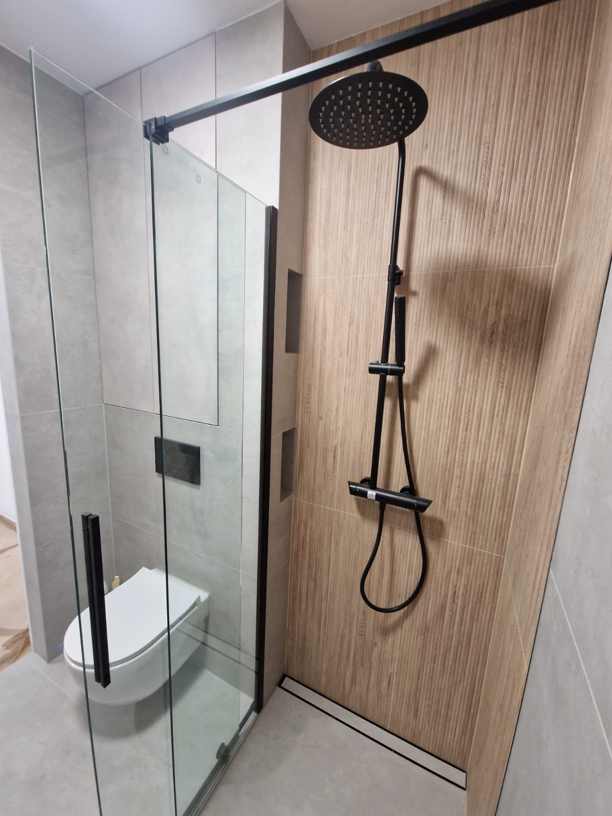 Nowoczesna łazienka z kabiną prysznicową typu walk-in, wykończona płytkami w odcieniach szarości i drewna, z czarną armaturą i minimalistyczną toaletą podwieszaną.