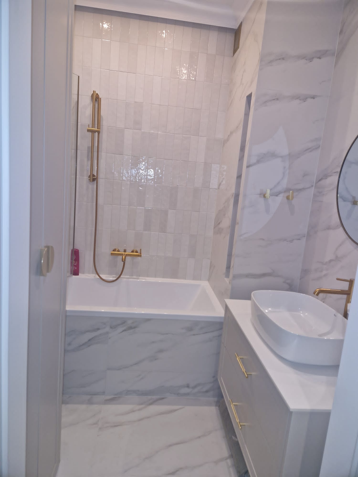 Nowoczesna łazienka z wanną obudowaną marmurem, białą umywalką nablatową i złotą armaturą. Ściany wyłożone białymi płytkami i marmurem.