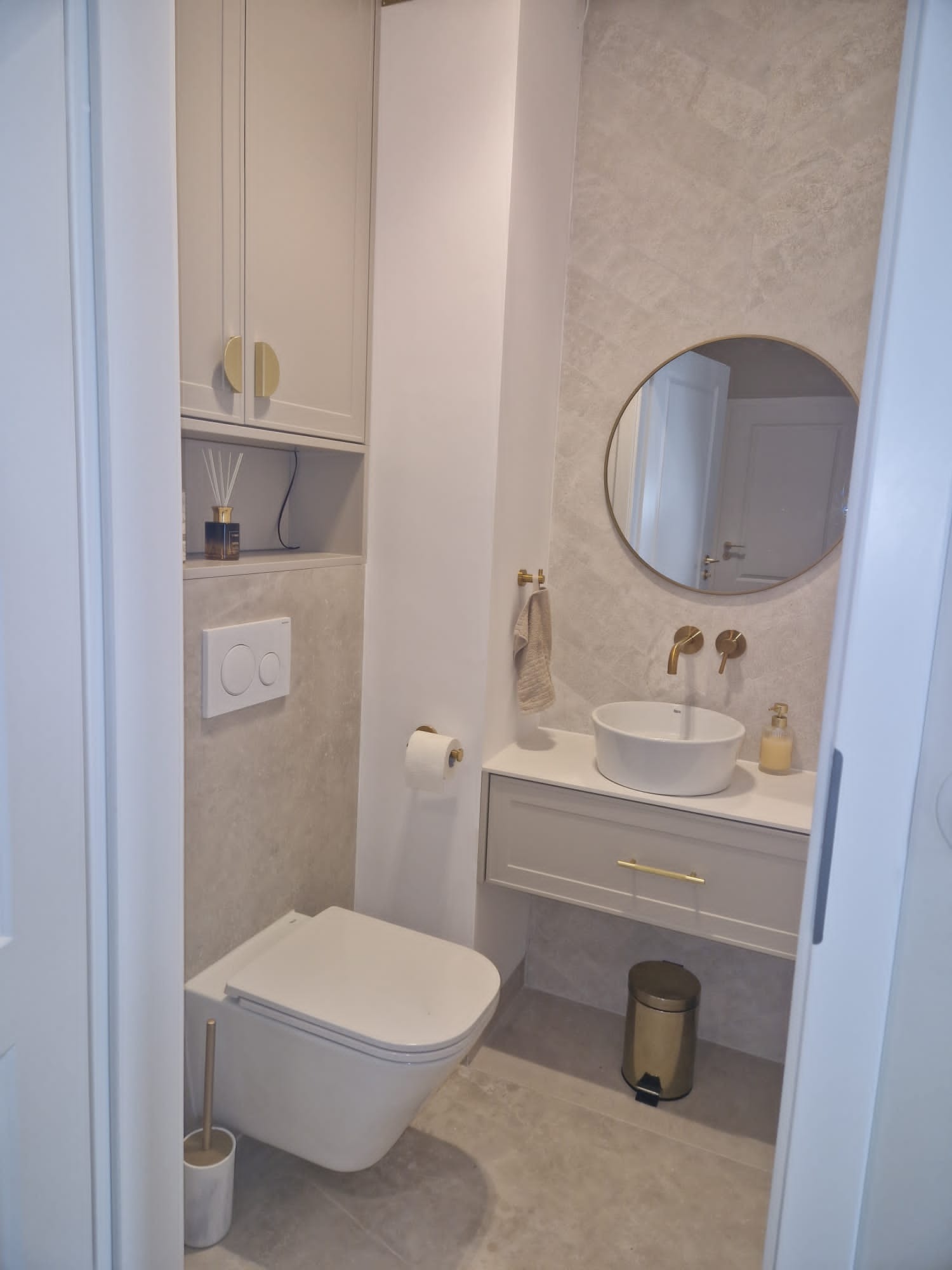 Jasna łazienka w odcieniach szarości z elementami złota: okrągłe lustro, bateria umywalkowa, uchwyt na ręcznik i kosz na śmieci. Umywalka nablatowa, toaleta podwieszana.