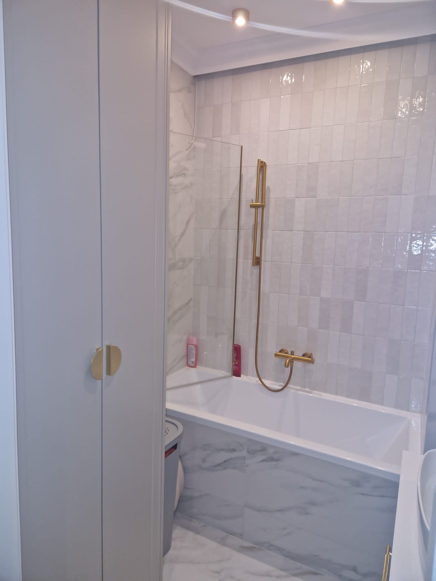 Wykończona łazienka w bieli i szarości z wanną, złotą armaturą i szafką z okrągłymi uchwytami. Marmurowe płytki na ścianach i podłodze. Nowoczesny design.