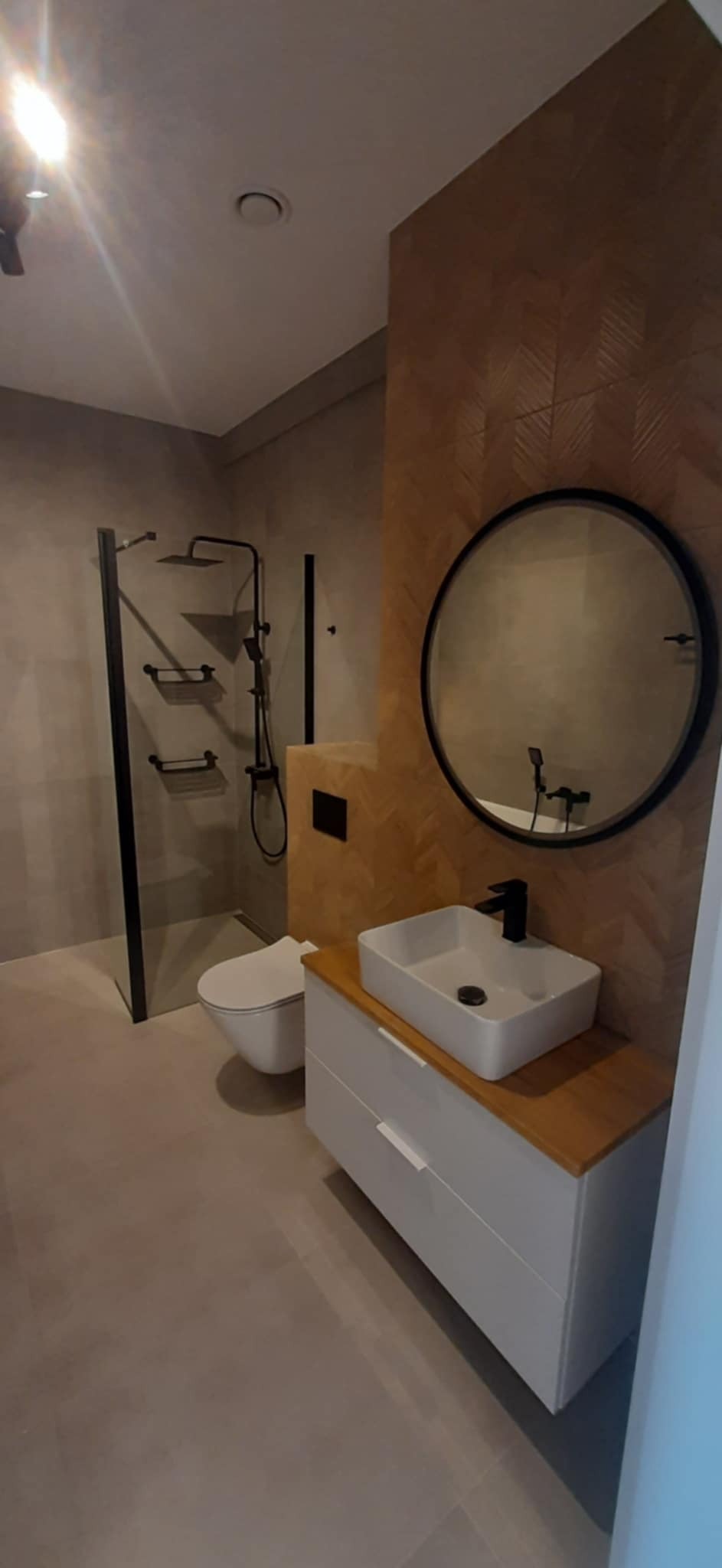 Nowoczesna łazienka z kabiną prysznicową typu walk-in, okrągłym lustrem i umywalką nablatową na szafce z drewnianym blatem. Ściana wykończona drewnopodobną mozaiką.