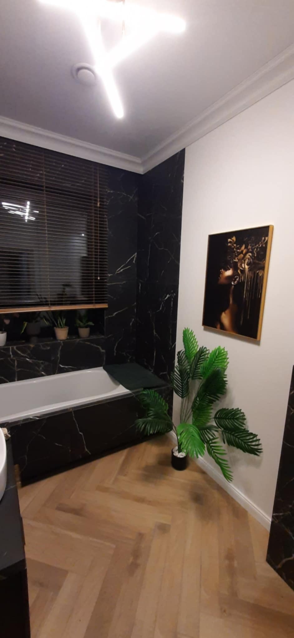 Elegancka łazienka z wanną obudowaną czarnym marmurem, drewnianą podłogą w jodełkę i obrazem na ścianie. Roślina doniczkowa dodaje świeżości.